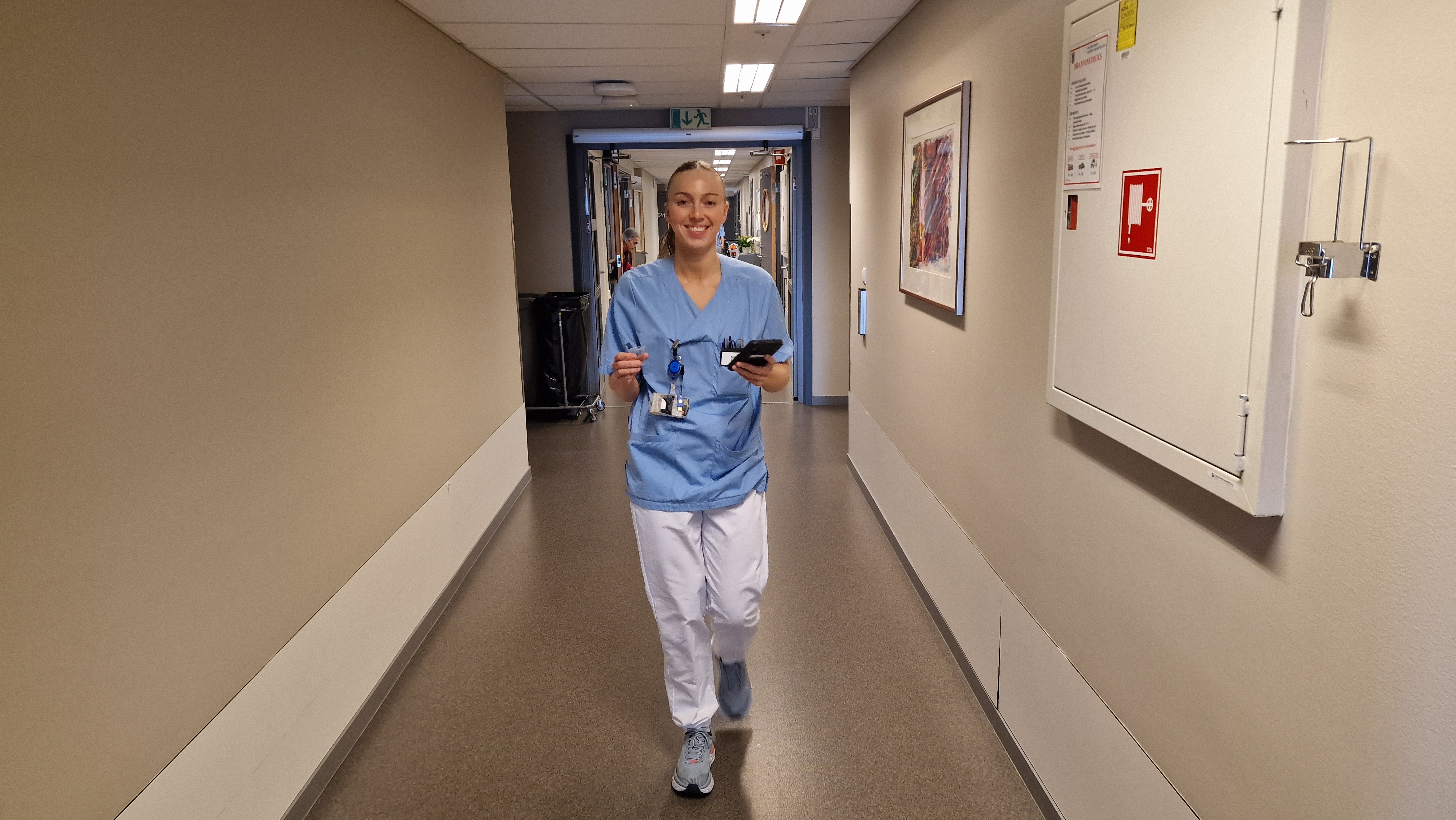 Sykepleier Maren Aslaksen går i en sykehuskorridor med mobilen i hånden.
