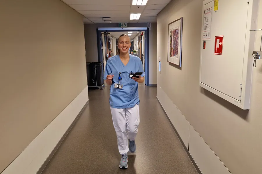 Sykepleier Maren Aslaksen går i en sykehuskorridor med mobilen i hånden.
