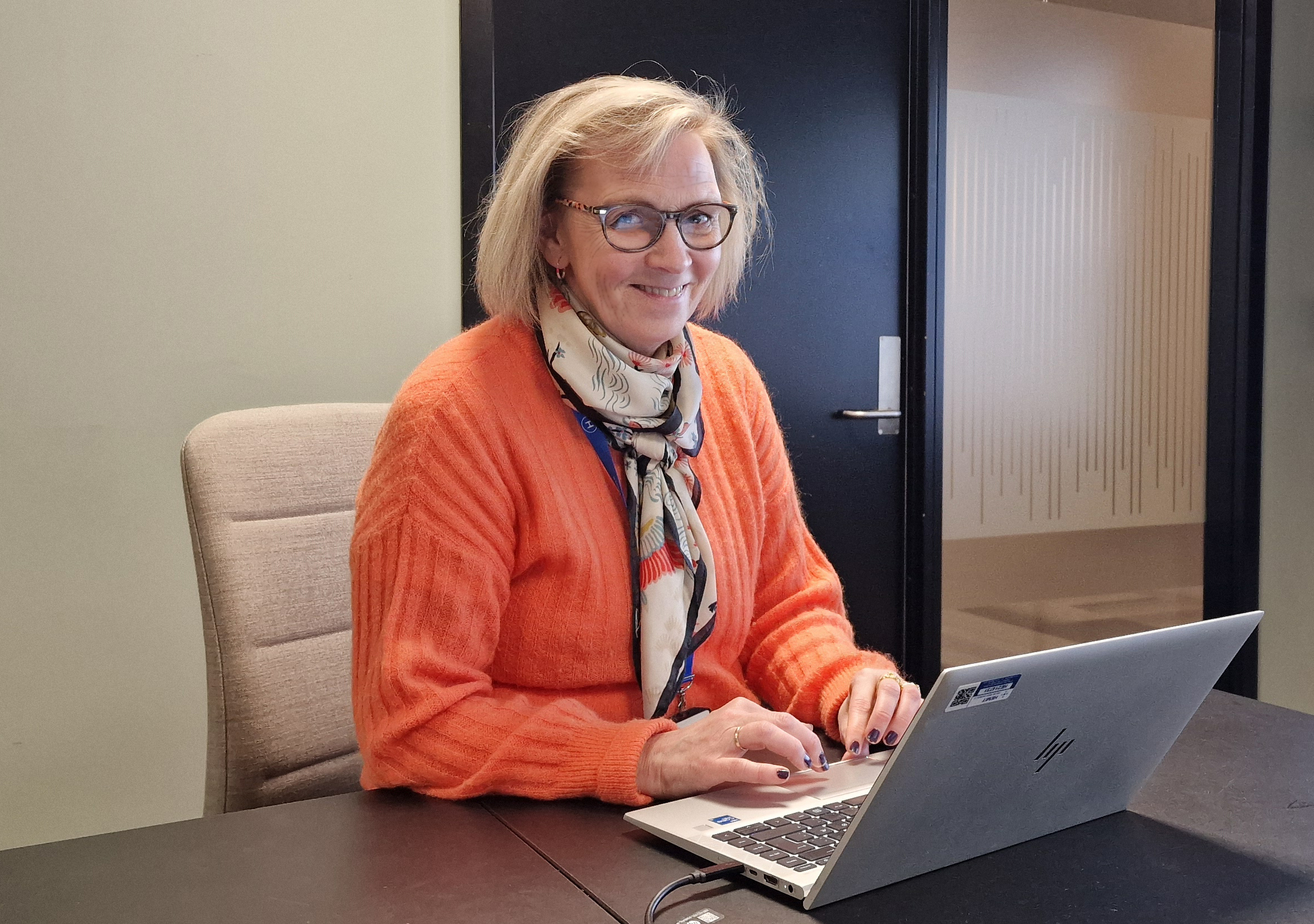 Hilde Grimnes Olsen har på oransje genser og sitter ved et bord med en bærbar PC