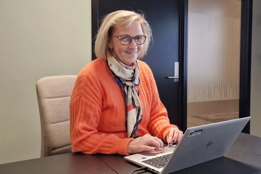 Hilde Grimnes Olsen har på oransje genser og sitter ved et bord med en bærbar PC